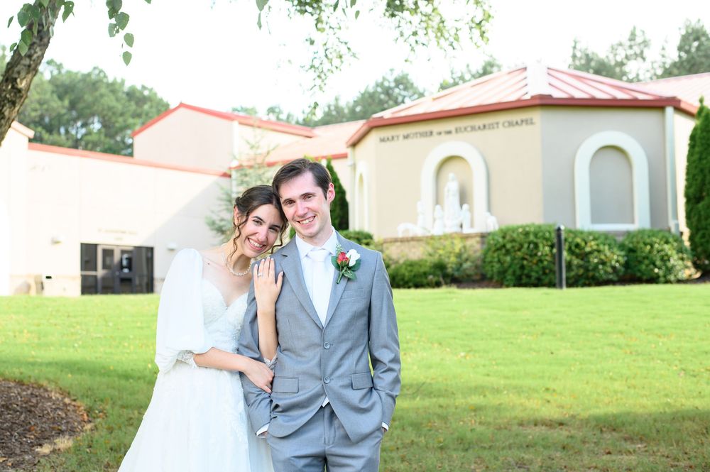 A romantic outdoor wedding portrait on a green lawn with a Mediterranean-style building in the background.