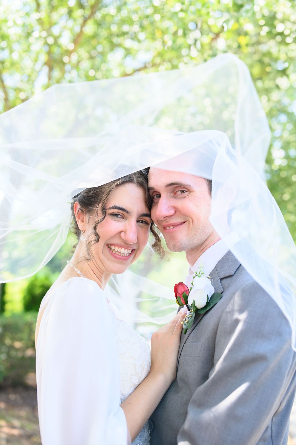 Bride and groom share an intimate moment under a flowing veil in a sunlit outdoor setting.
