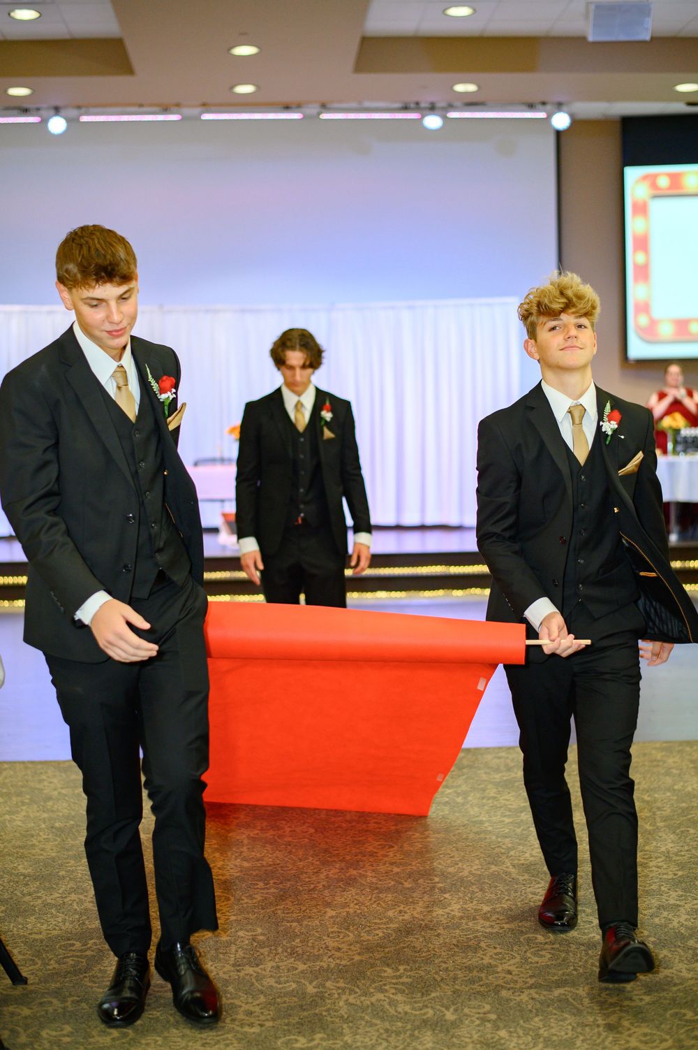 Young boys in formal black suits walk down a red carpet at an event.