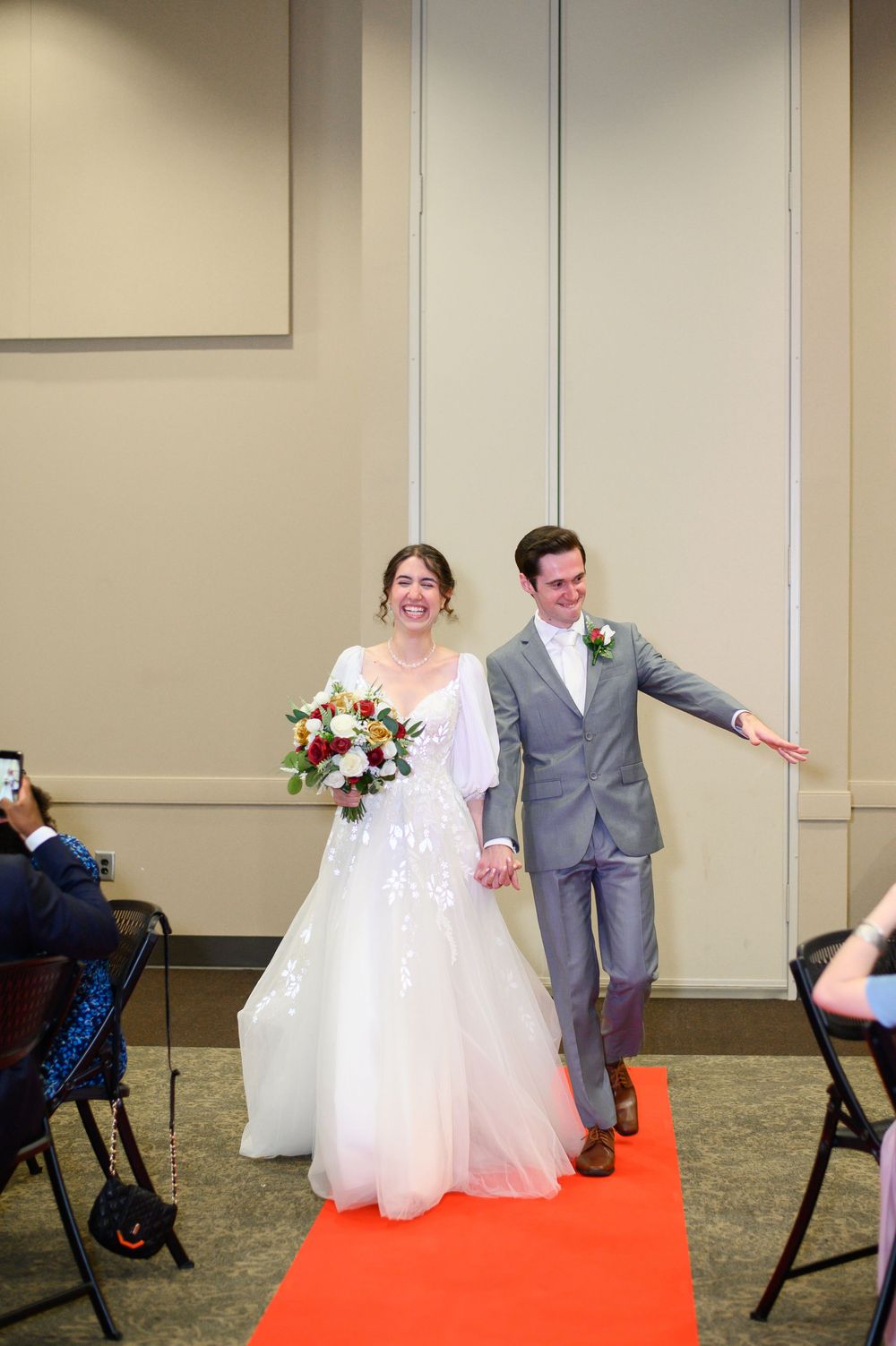 A happy couple walks down a red carpet aisle at their wedding ceremony, wearing grey suit and white gown.