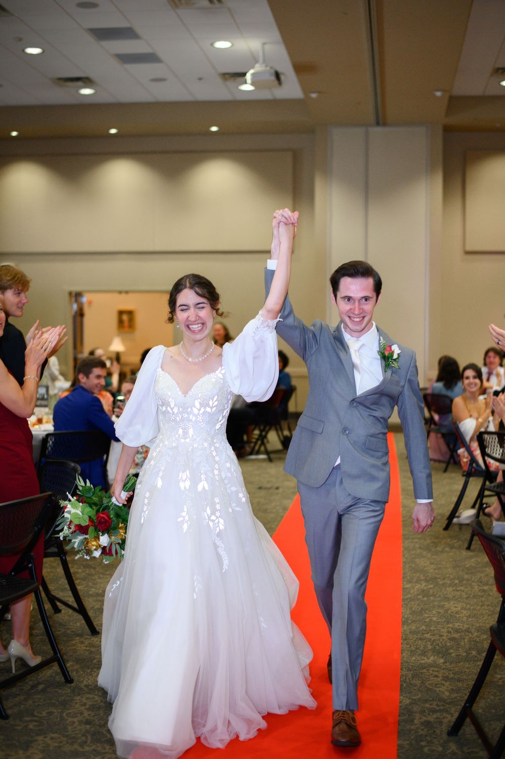 Wedding couple and guests dancing together in a reception hall with gray folding chairs.