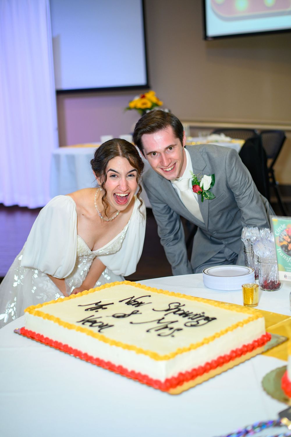 A joyful couple in wedding attire lean over their decorated reception cake, sharing a fun moment together.