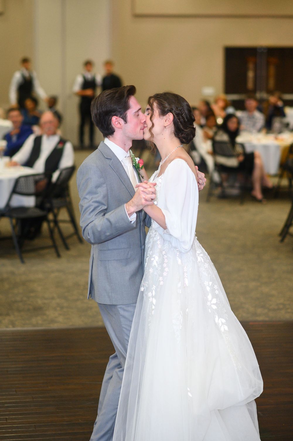 A couple shares their first dance as newlyweds in a reception hall with guests seated at tables in the background.