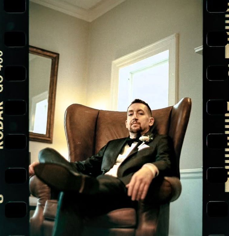 Person in suit sitting casually in brown leather chair against white wall. 35 mm Film Wedding Photography Pittsburgh Pennsylvania.
