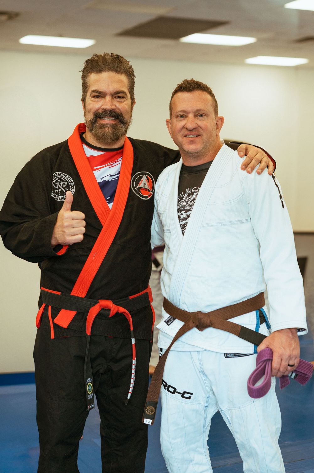 Two martial artists in Brazilian Jiu-Jitsu gis posing together at a training facility with fluorescent lighting overhead.