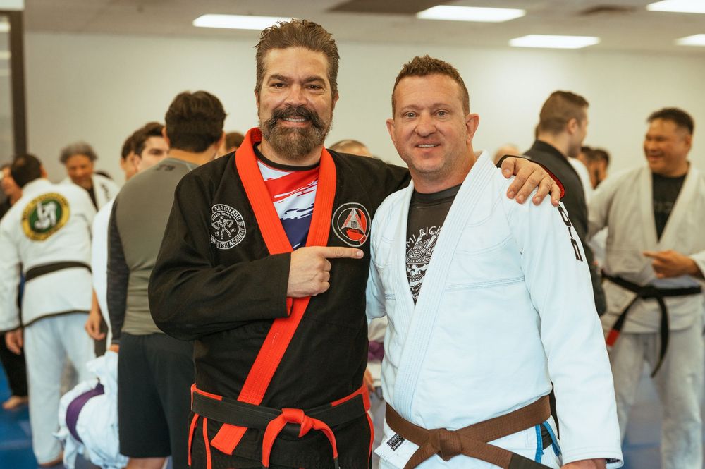 Two practitioners in martial arts uniforms pose together at a training facility with others visible in the background.