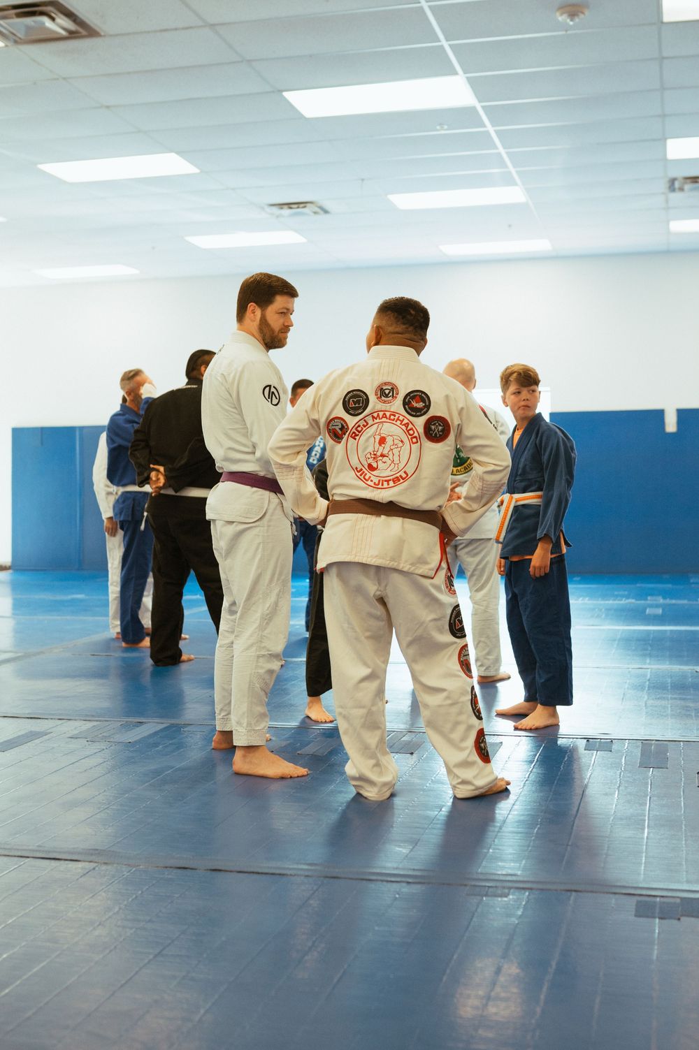 Students practicing martial arts in white uniforms on blue mats in a bright indoor training facility.