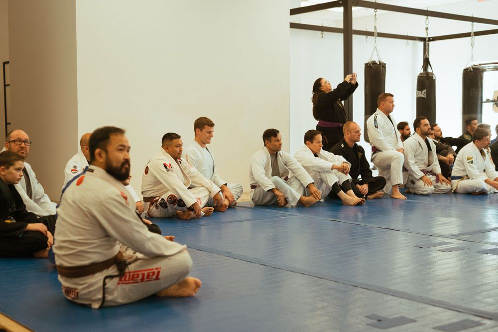 Students in white martial arts uniforms sit attentively on blue mats during training at a mixed martial arts studio.