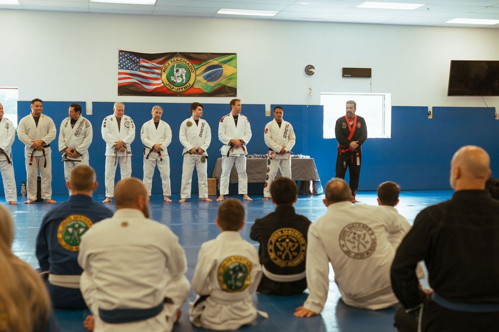 Students in white martial arts uniforms bow in respect at a training session with flags and certificates on display.