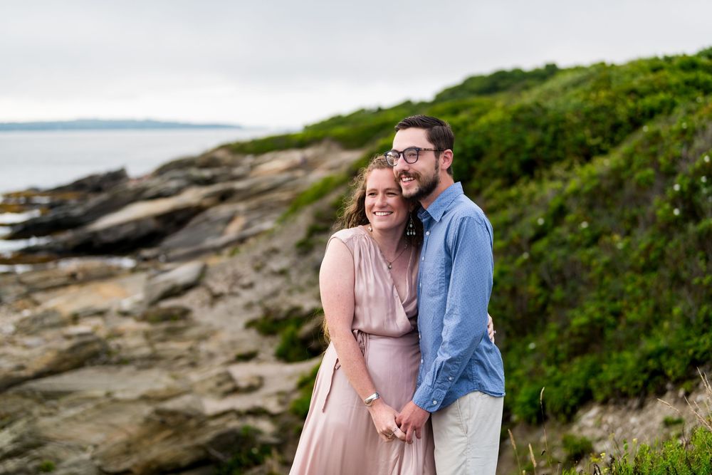 A couple in light blue and pink clothing embraces on a rocky coastal cliff during sunset.