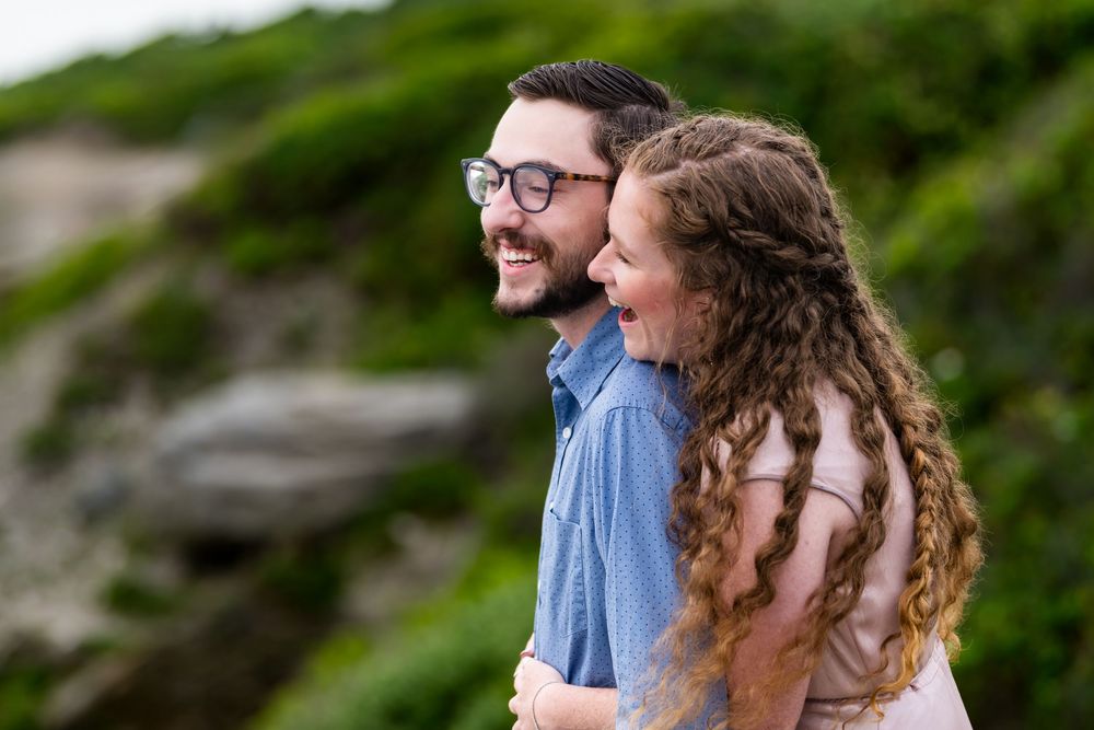 A joyful couple in casual attire share a laugh together outdoors with a blurred natural background.