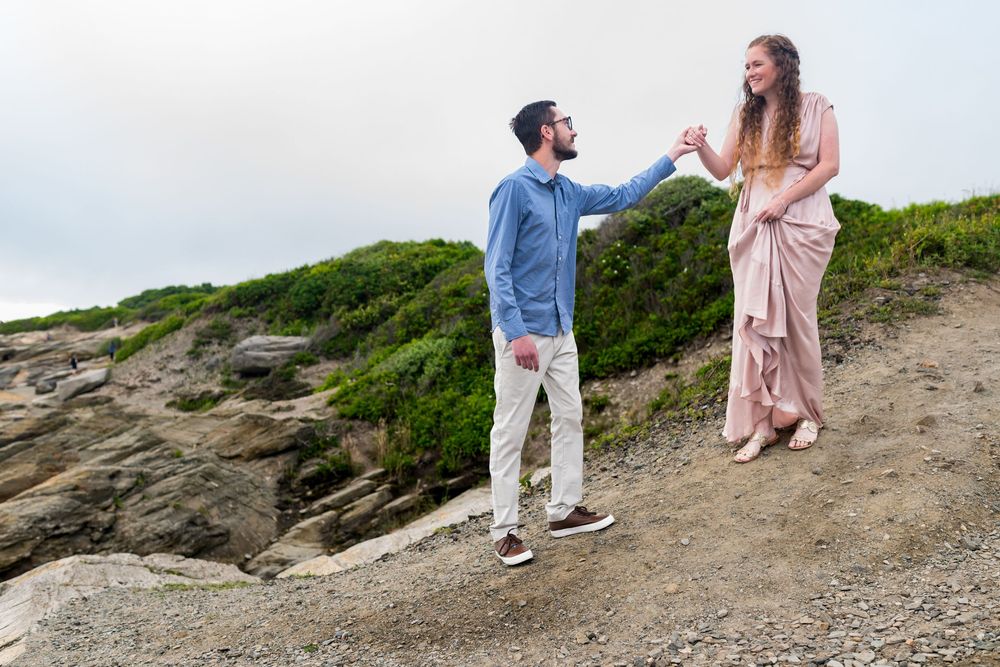 A couple walks along a scenic coastal path with green shrubs and rocky terrain in golden hour lighting.