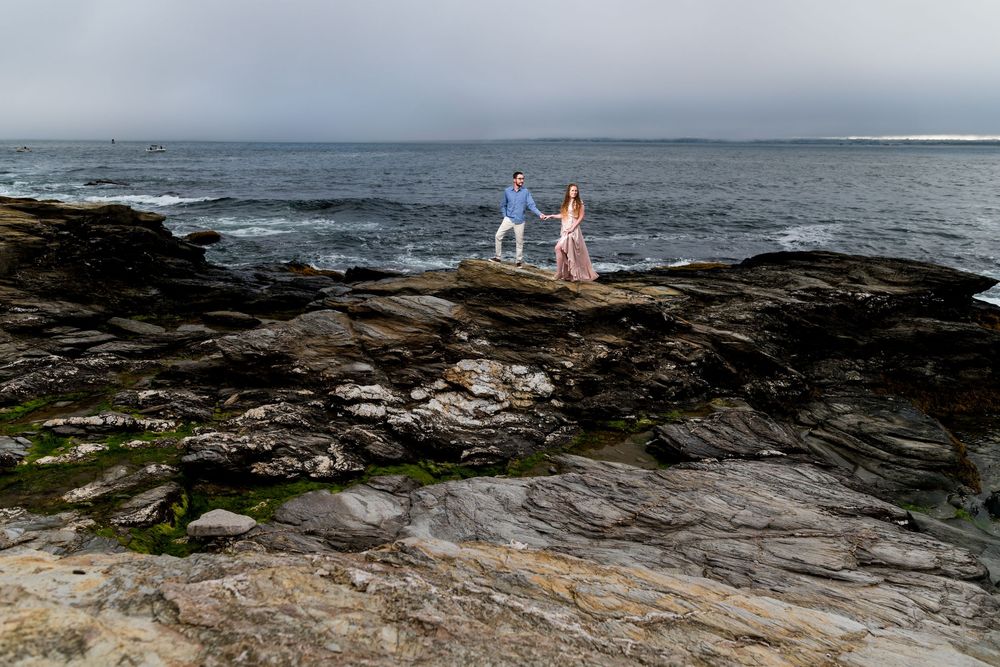 Two figures stand on a rugged coastal shoreline as waves crash against the rocky Maine coast during sunset.