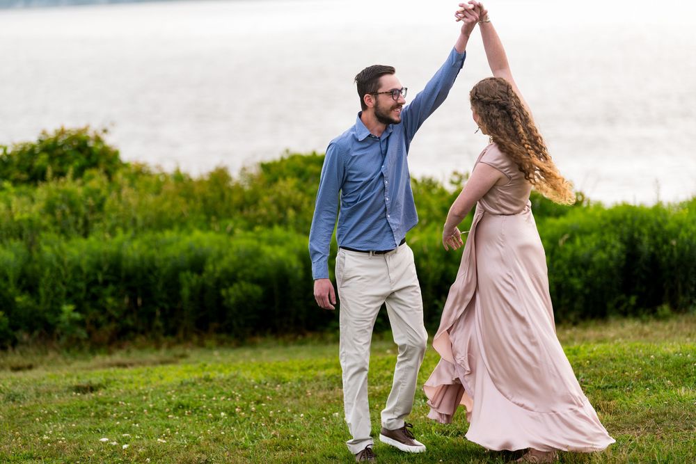 A couple dances together on a grassy hill during an overcast day, with her pink dress flowing in the breeze.