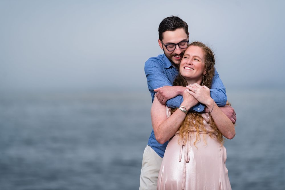 A loving couple embraces by the ocean, with a misty gray seascape in the background.