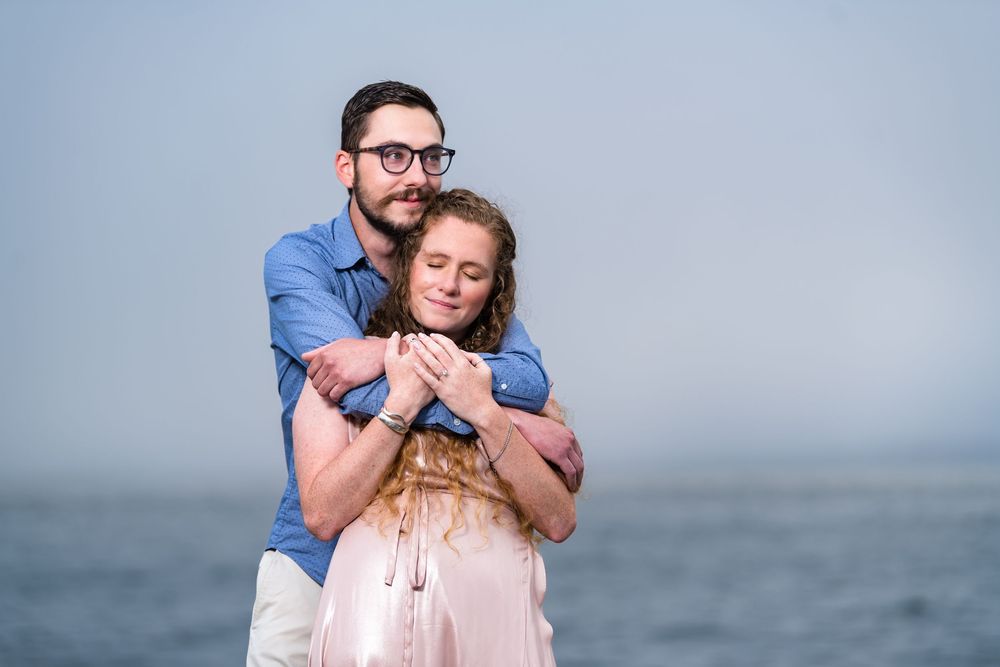 Couple in blue and pink outfits embrace each other while enjoying a romantic moment at the beach during sunset.