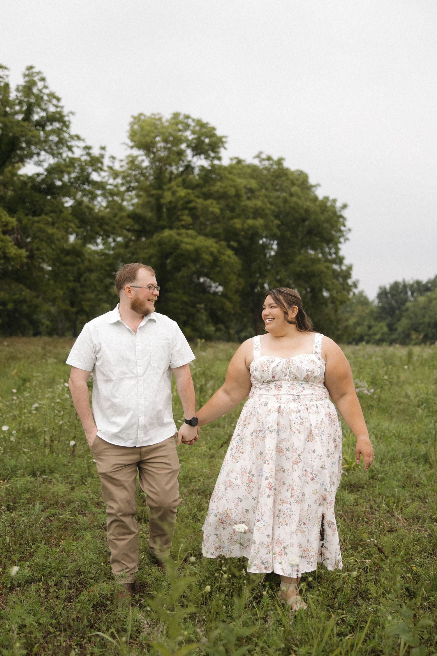 A couple walks together through a green summer meadow, she in a floral dress and he in a white shirt and khakis.