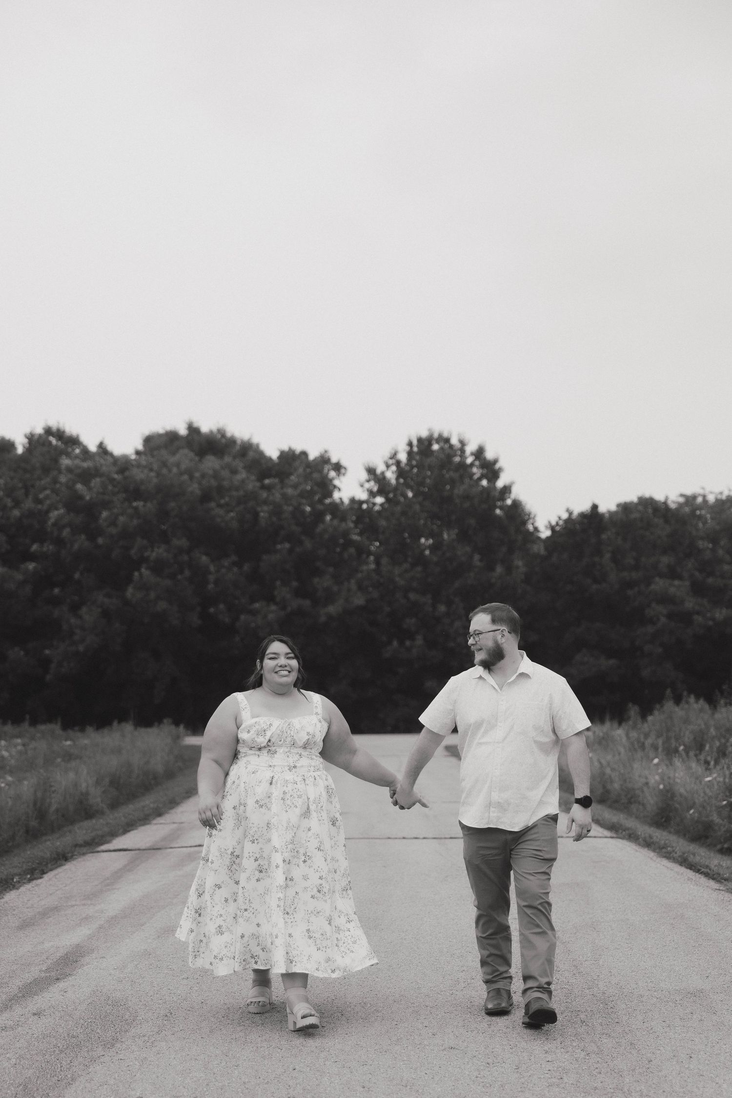 A romantic black and white photo of a couple holding hands while walking down a rural road on a summer day.