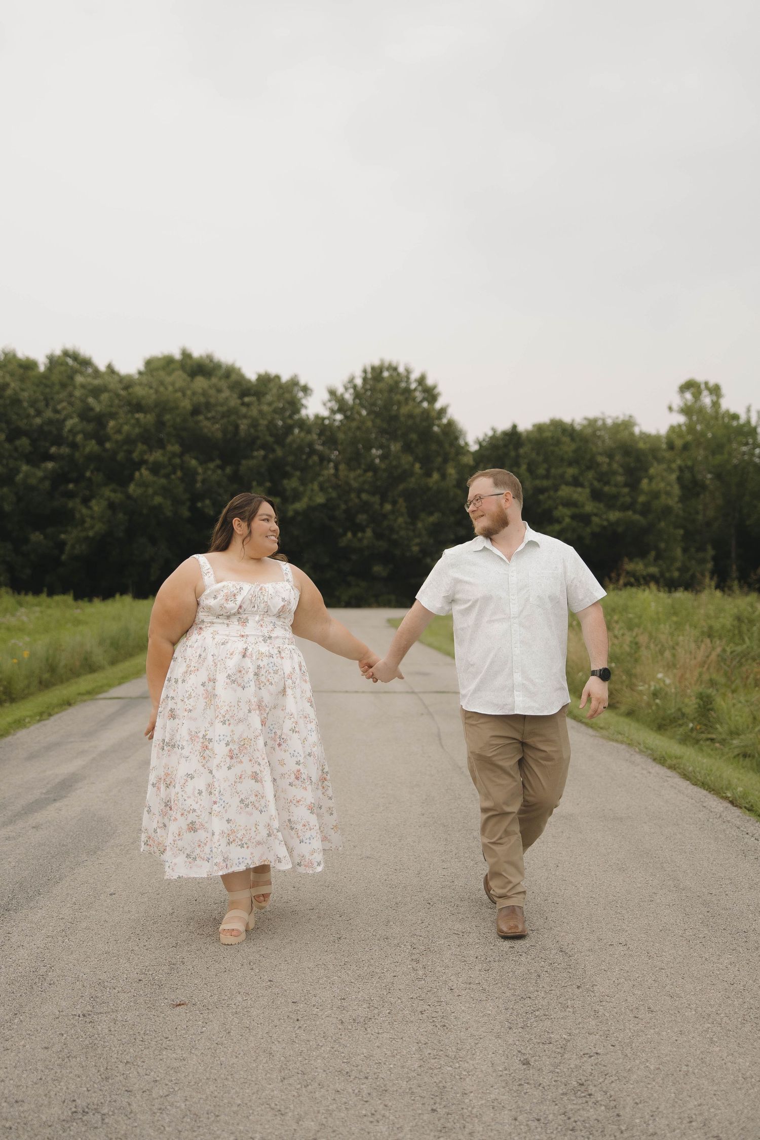 A couple in white and khaki clothing walks on a gravel path surrounded by green fields and trees.