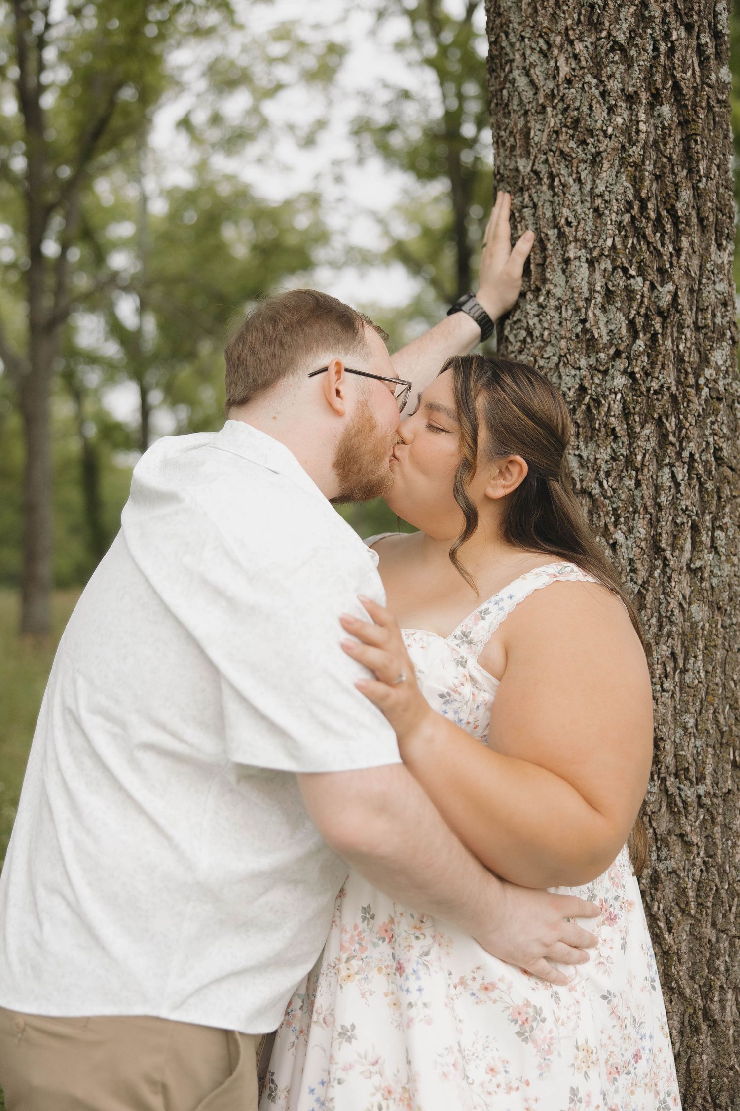 A romantic outdoor photoshoot shows a couple sharing tender kisses against tree trunks in a woodland setting.
