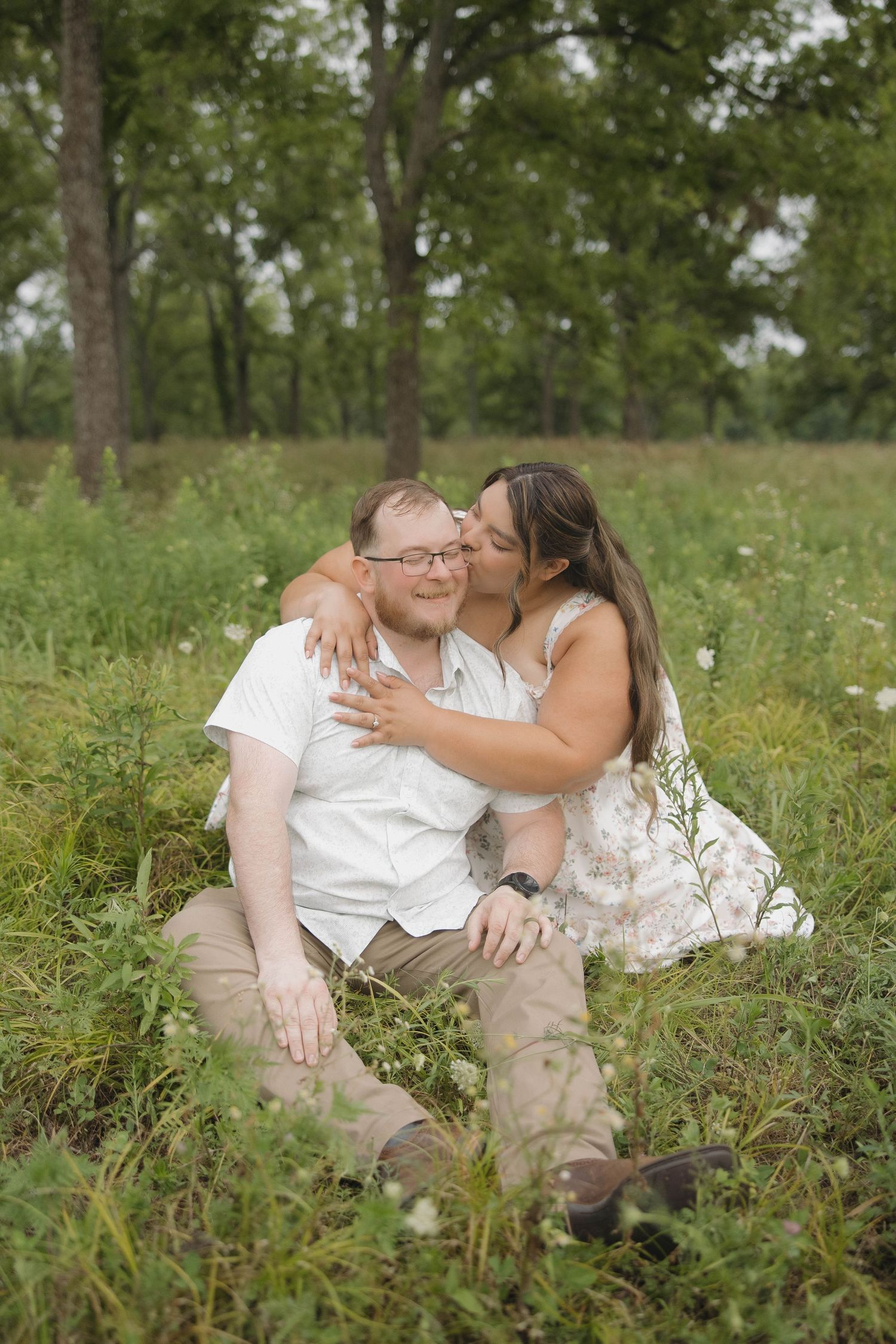 Intimate couple sharing tender moment together while sitting in a grassy meadow surrounded by trees on summer day.