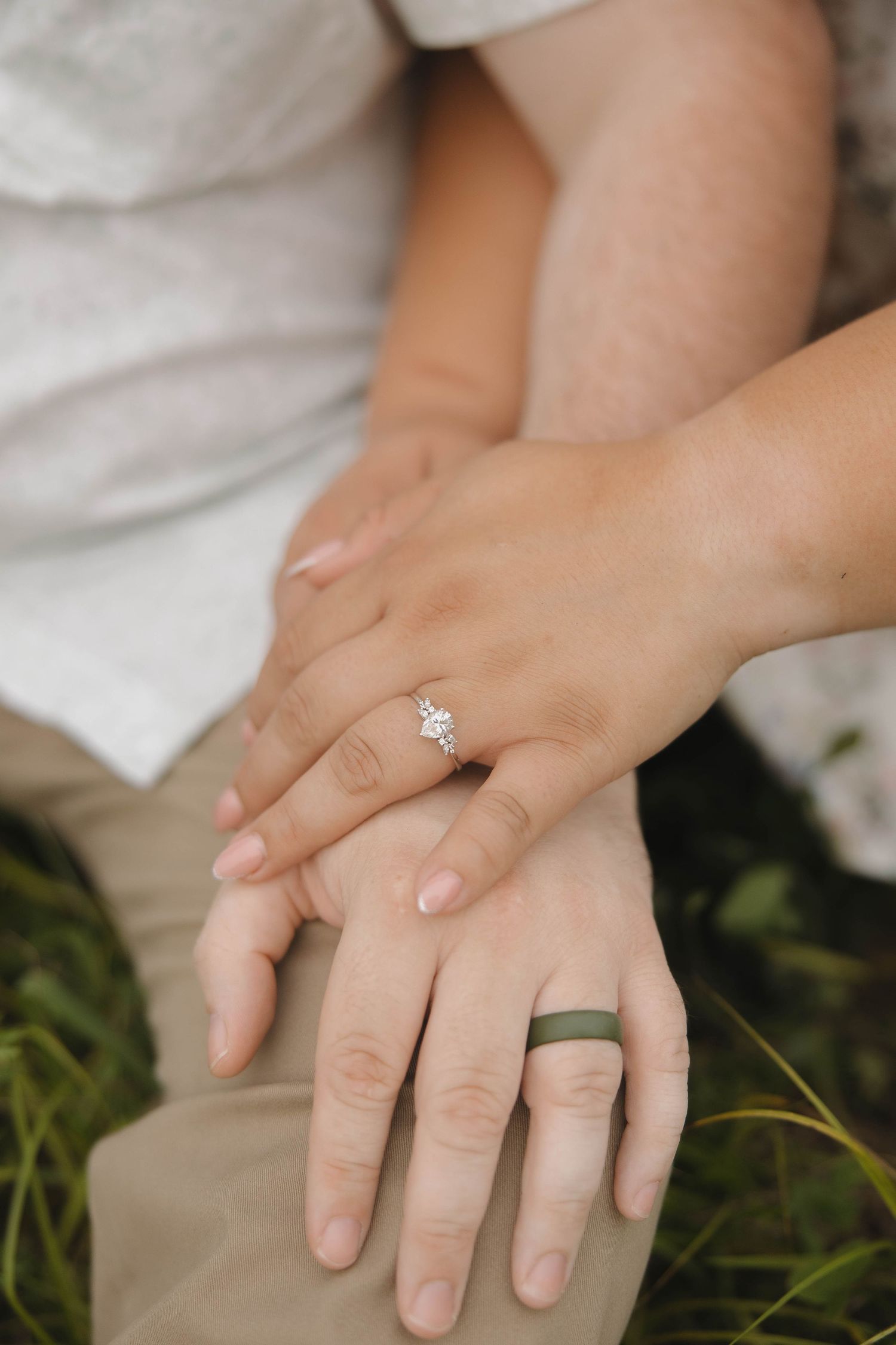 Close-up of hands wearing engagement ring and wedding band resting on white fabric with grass beneath.