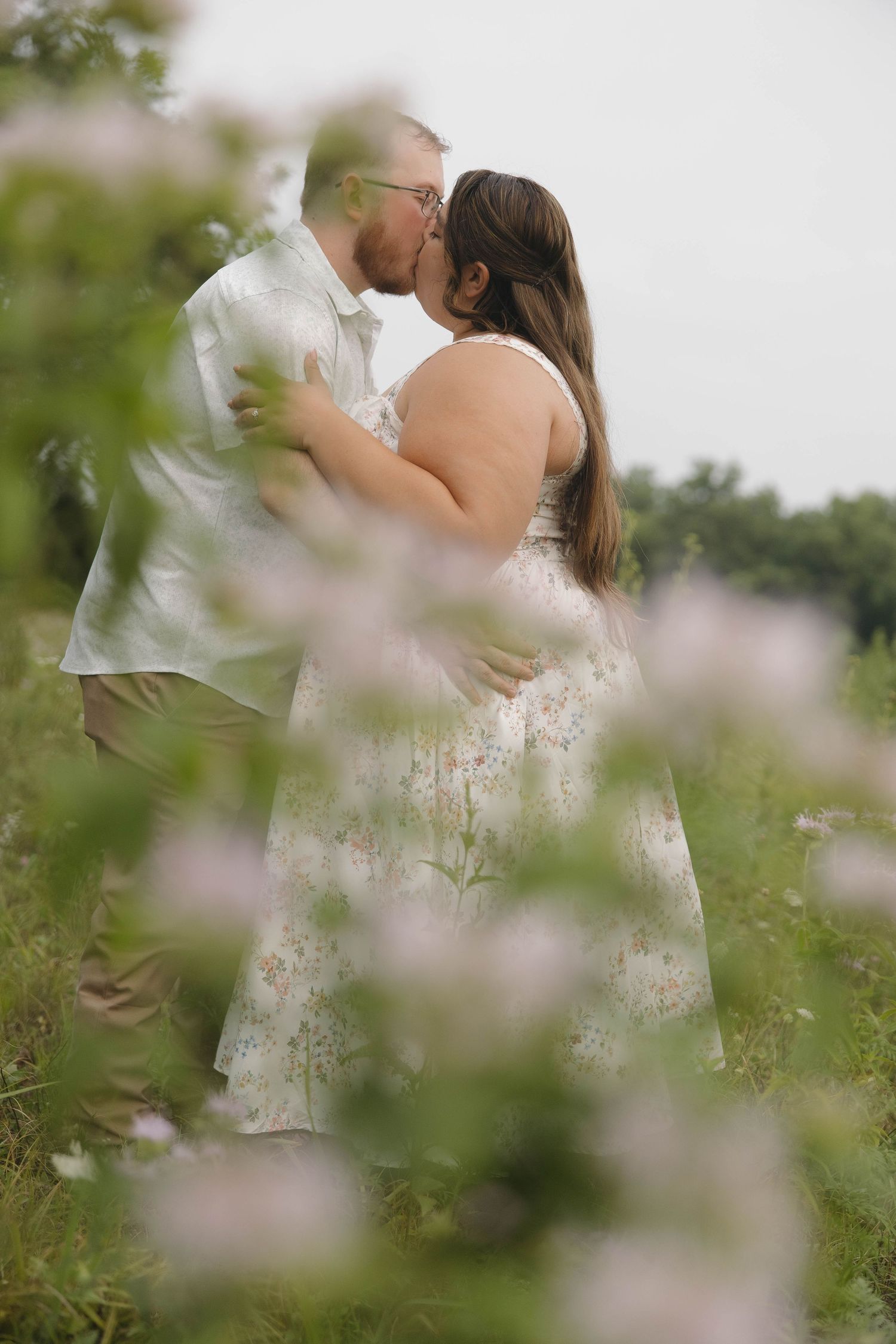 A romantic couple embraces in a meadow surrounded by soft white wildflowers on an overcast day.