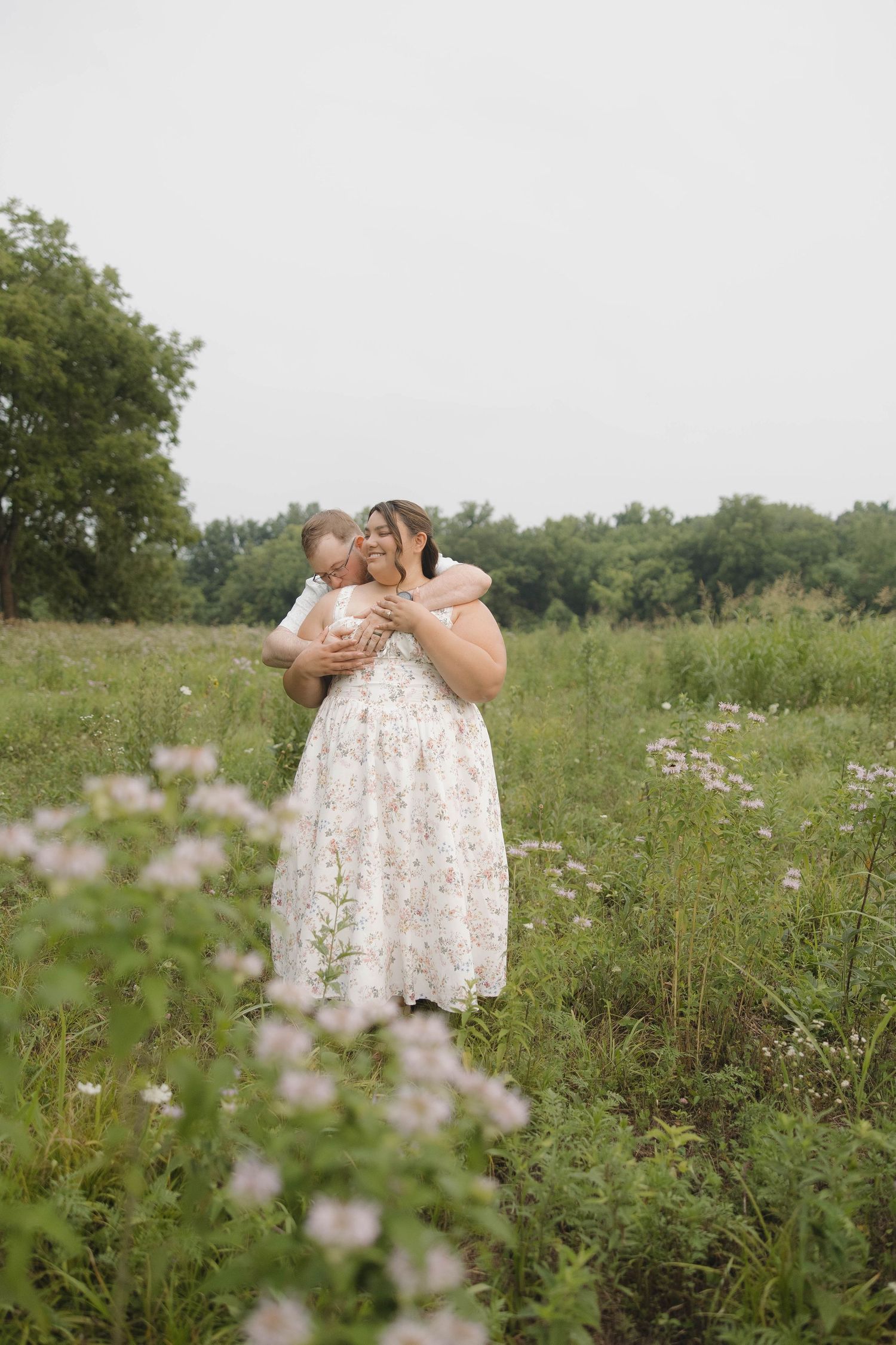 A couple standing in a wildflower meadow wearing a floral white dress share an intimate moment on a cloudy day.