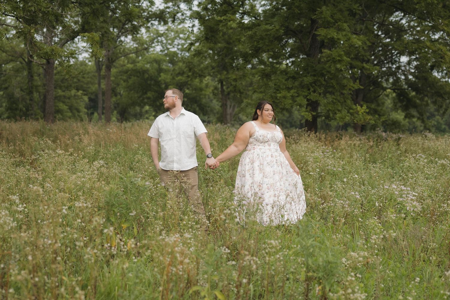 A couple sits in a wildflower meadow surrounded by lush green trees on a summer day.