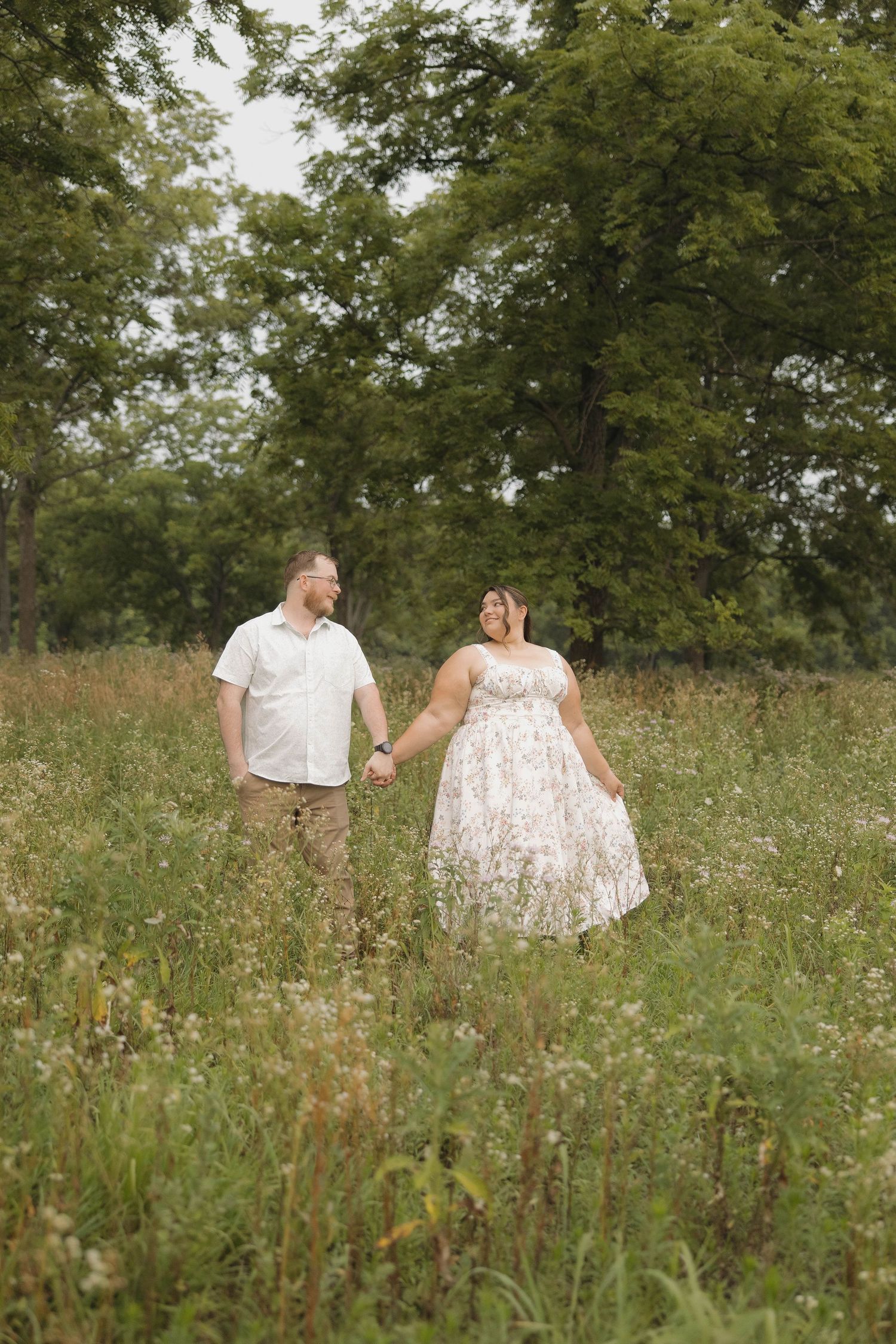 A couple holding hands while walking through a wildflower meadow on a summer day, surrounded by tall trees.
