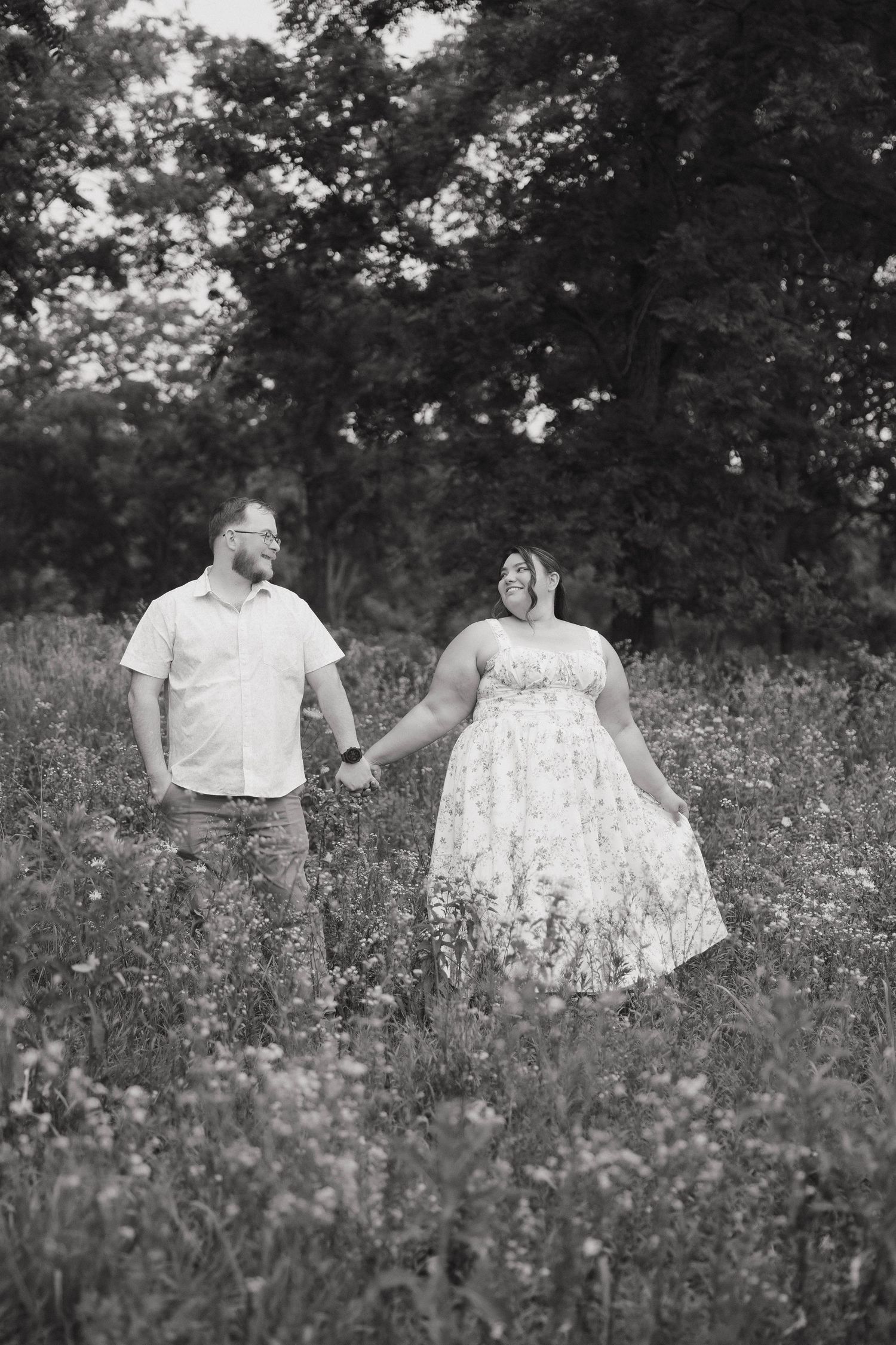 A couple holds hands while standing in a wildflower meadow, captured in black and white photography.
