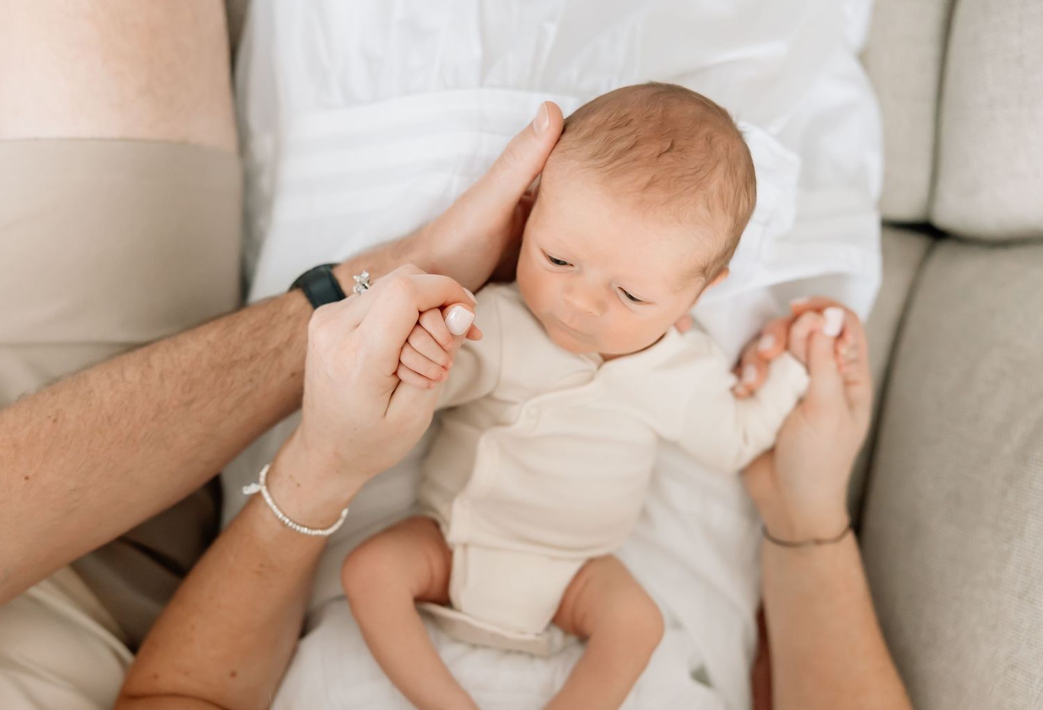 A peaceful sleeping baby in white clothing holds an adult's hand while resting on soft fabric.