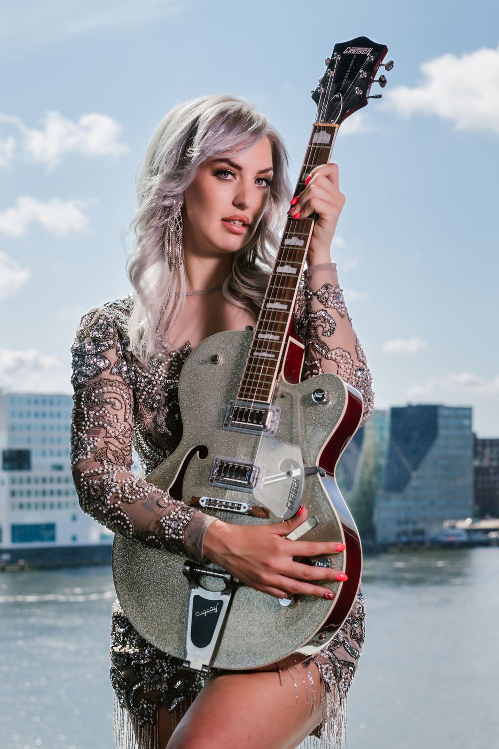 Fashion model poses with silver guitar against city skyline and water backdrop in glamorous sparkly outfit.