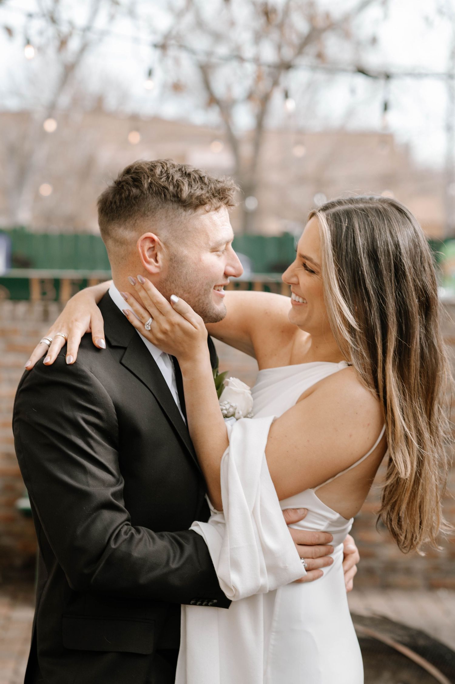 A romantic wedding couple shares an intimate moment outdoors with blurred lights and trees in the background.