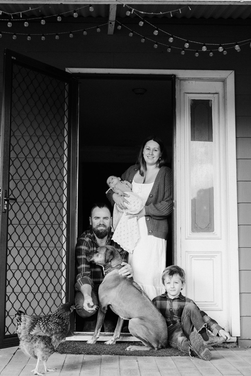 A black and white photo of a family with pets including a dog and chicken on their front porch with string lights overhead.
