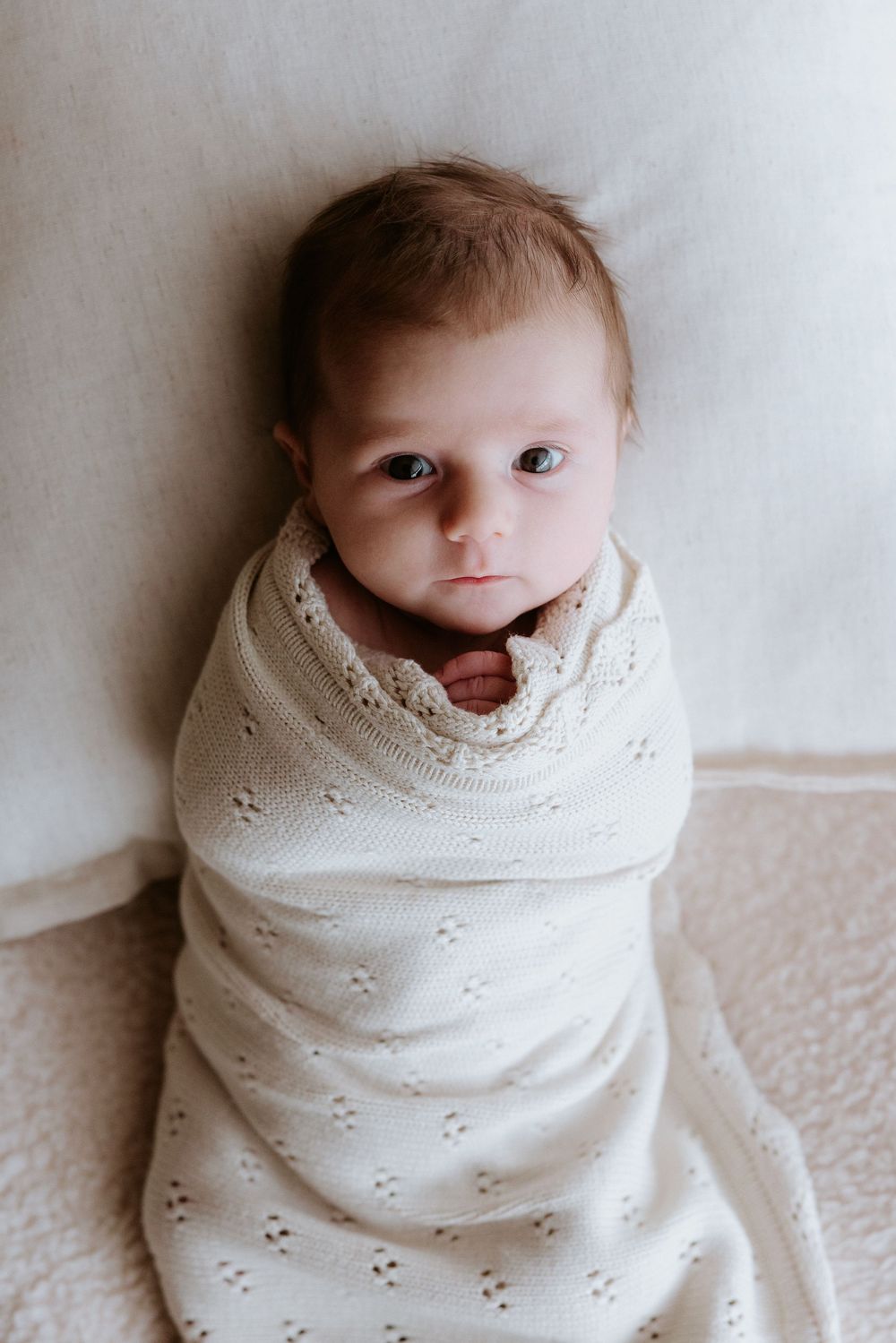 A tiny baby wrapped in a soft white eyelet blanket lies against a light background looking at the camera.