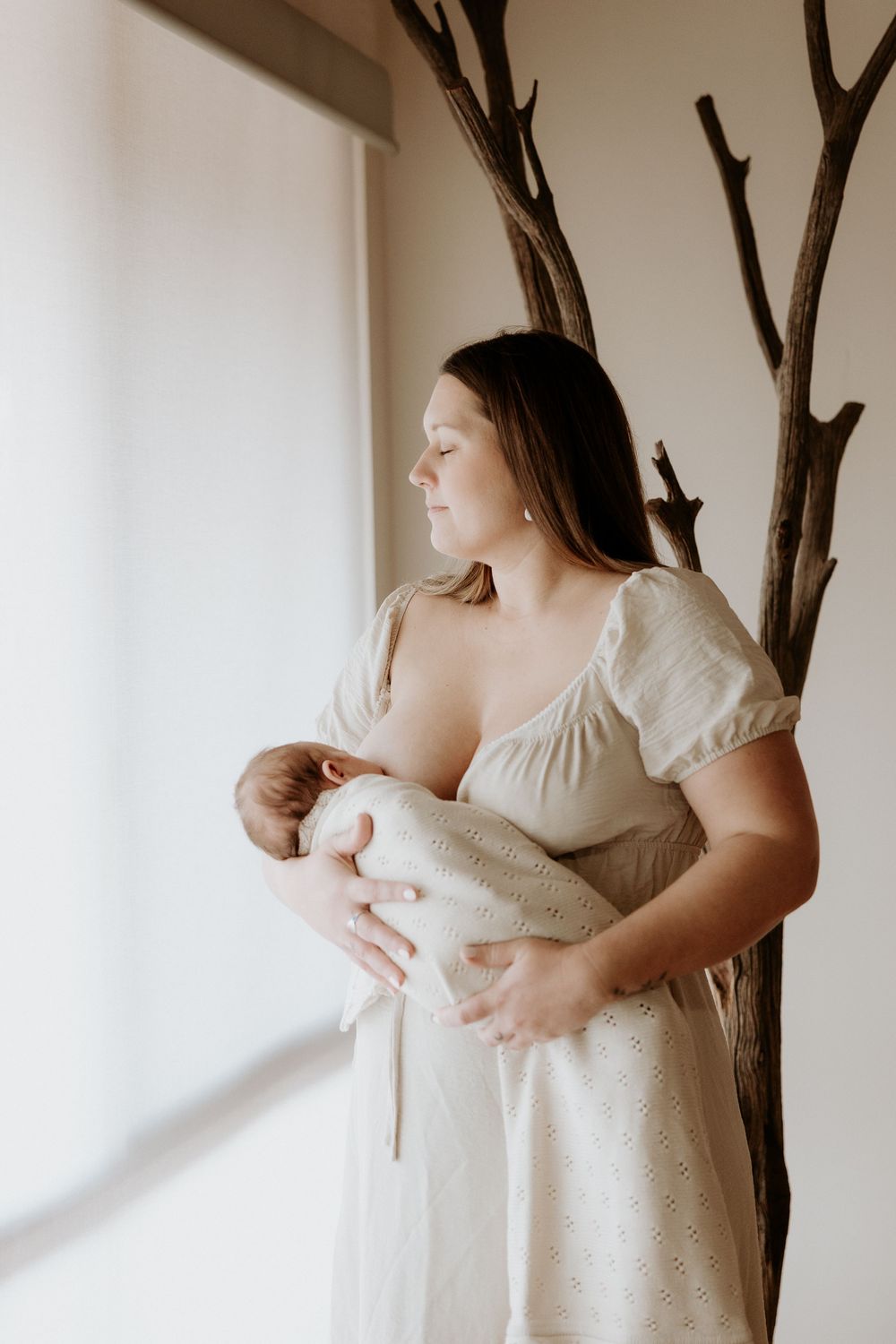 A mother in a white dress cradles her newborn baby against a soft white background while sitting near branches.