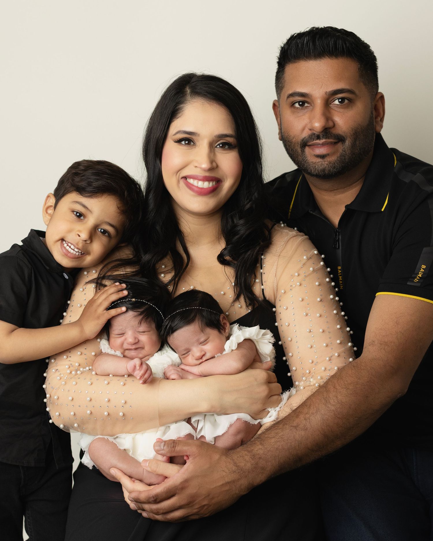 Mom holds twin newborns in tiny white dresses while her son places a hand and dad snuggles in