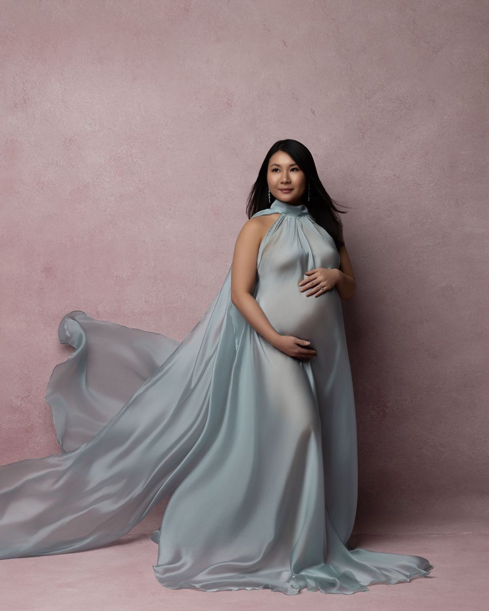 A flowing light blue maternity gown with elegant draping fabric captures movement in a studio setting with pink backdrop.