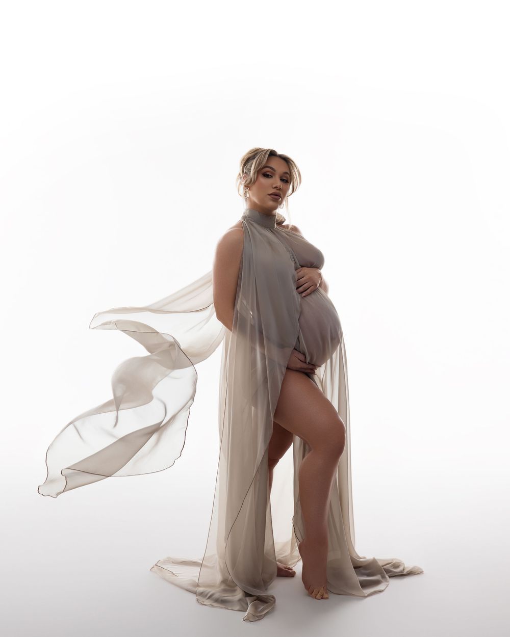 Elegant maternity photo featuring flowing sheer fabric draping around a pregnant figure against a bright white background.