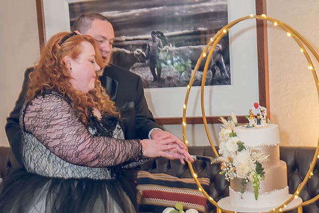 A couple cuts into their elegant three-tiered wedding cake decorated with white flowers beside a gold hoop display.