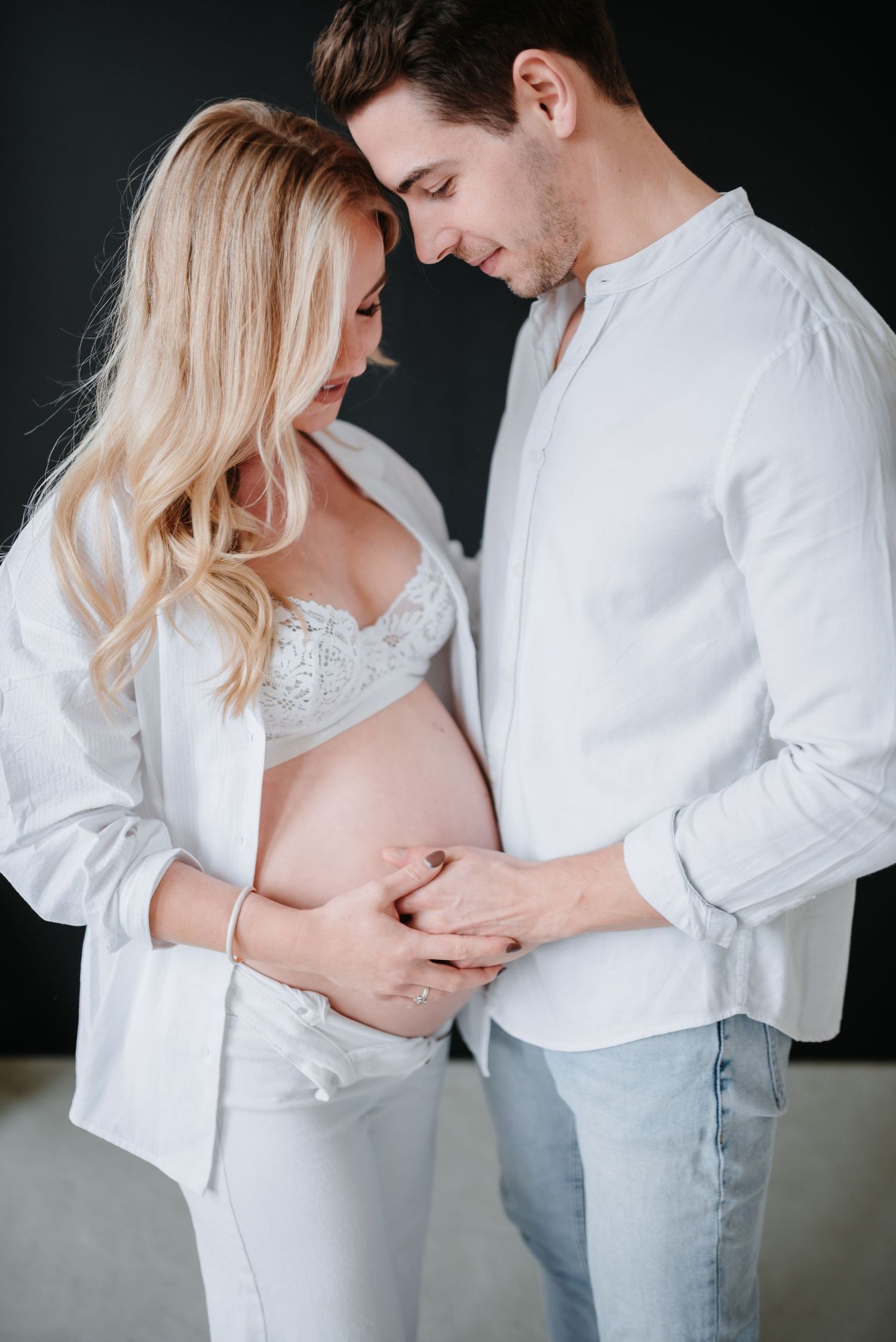 Romantic maternity photo of couple in white shirts touching pregnant belly against dark background.