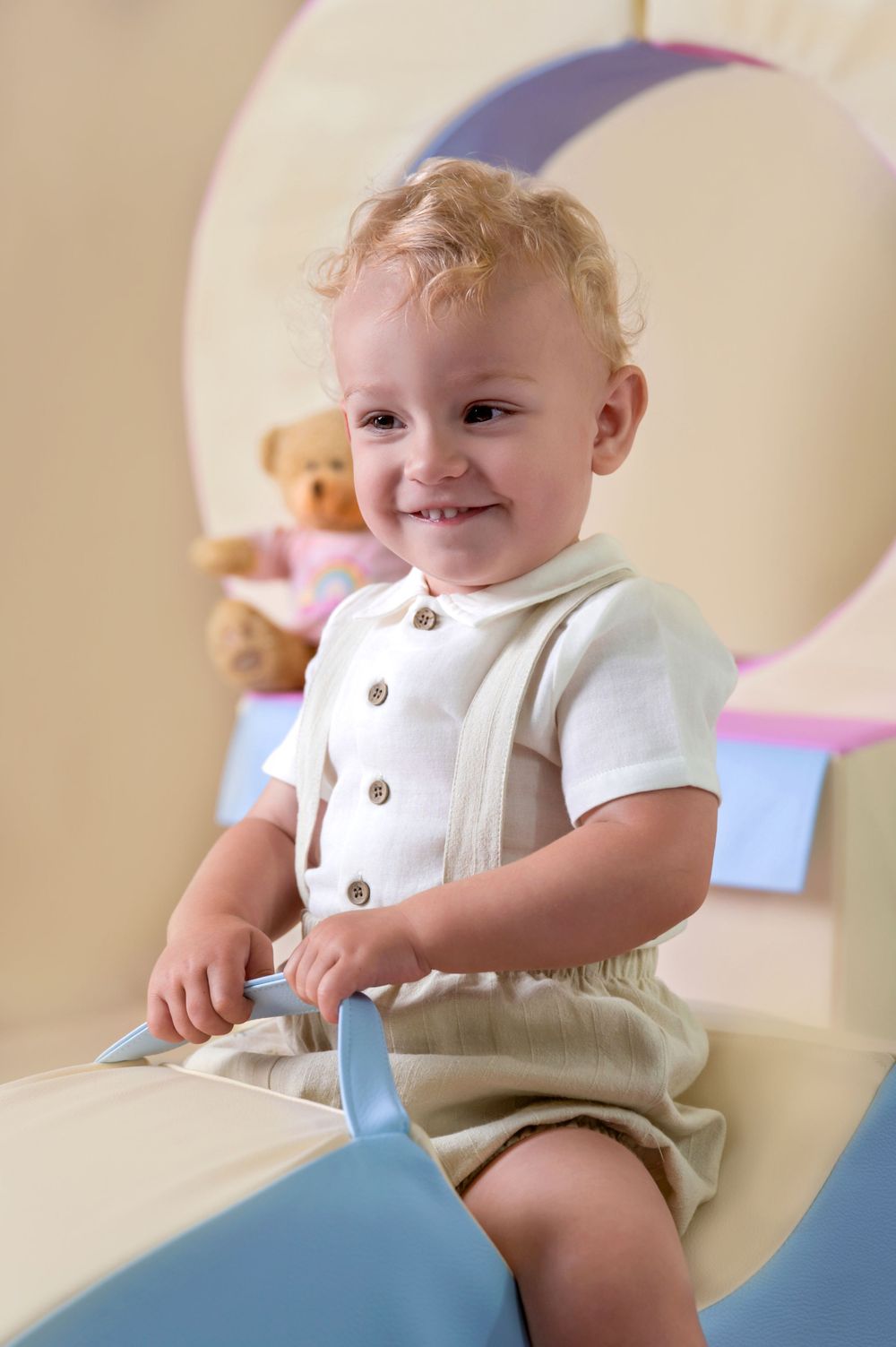 A cheerful toddler in a white shirt and beige shorts plays on a blue cushioned rocker with a teddy bear behind.