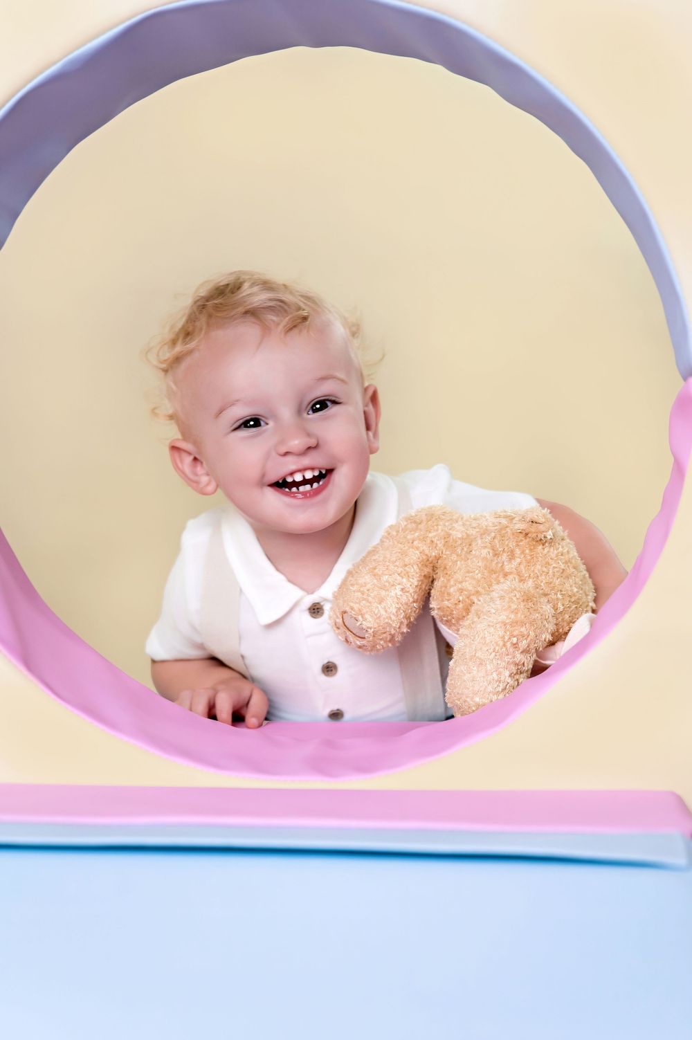 A happy toddler peeks through a circular frame while holding a plush teddy bear toy.