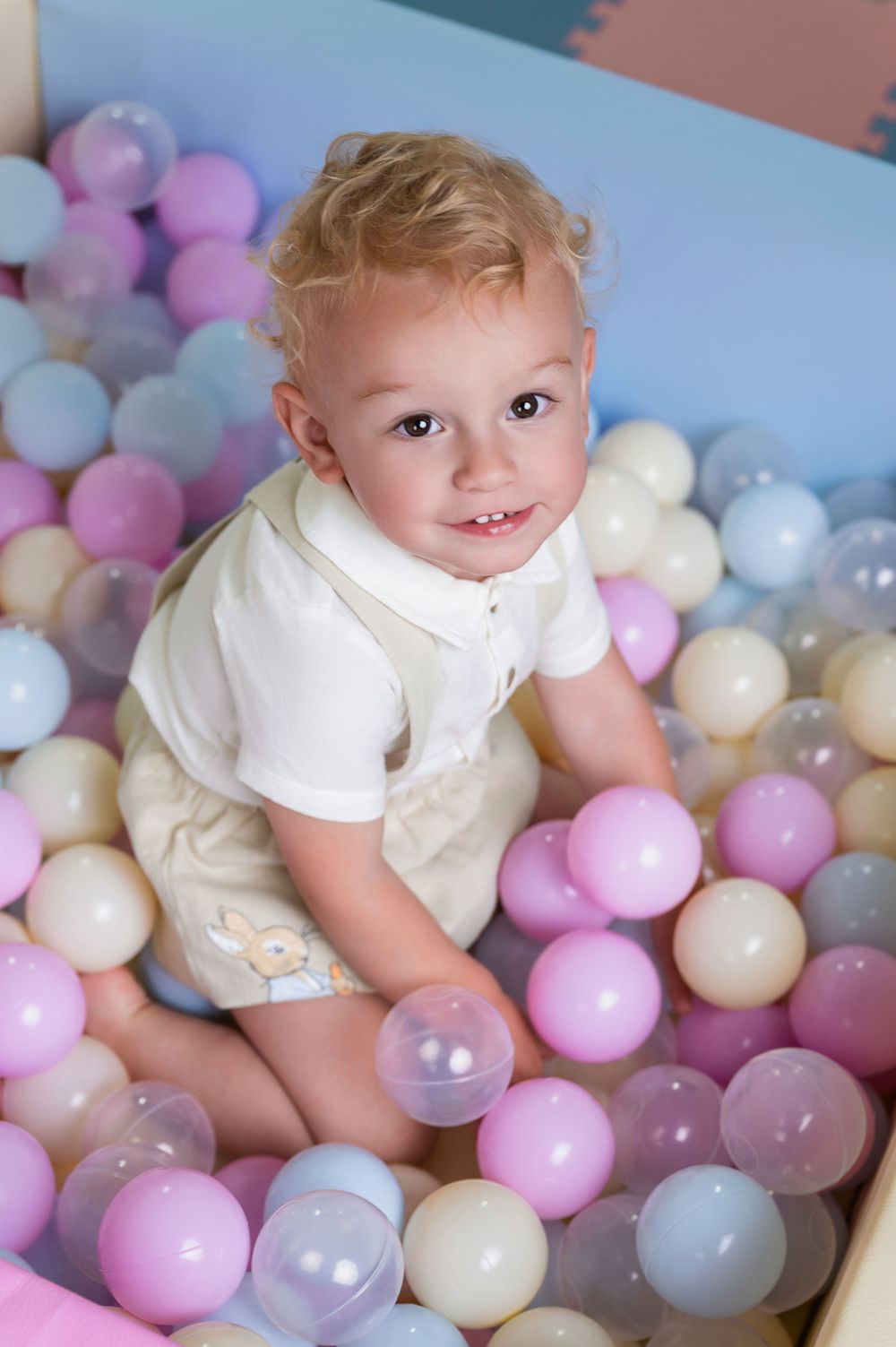A young child plays in a ball pit filled with pastel pink, blue and white plastic balls.