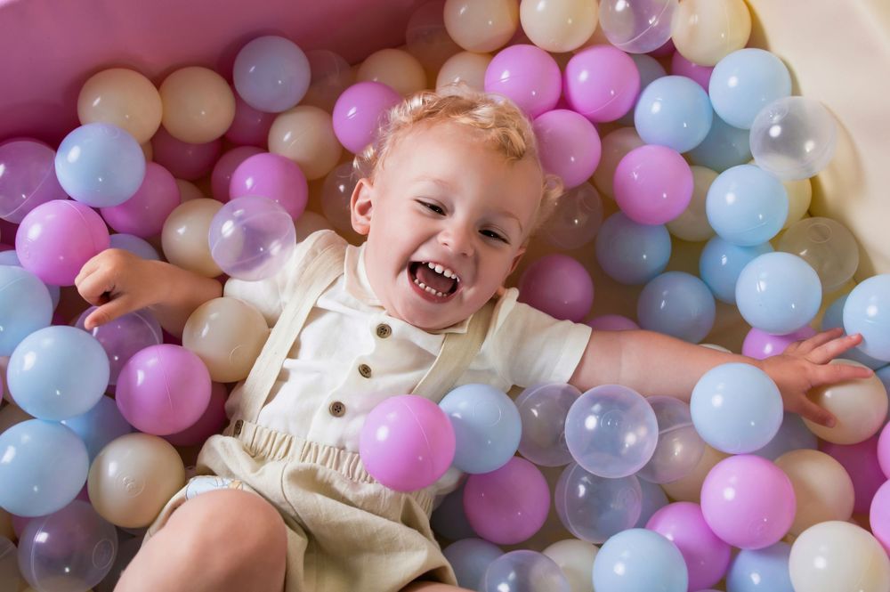 A laughing toddler lies in a ball pit filled with pastel blue, pink, and cream colored plastic spheres.