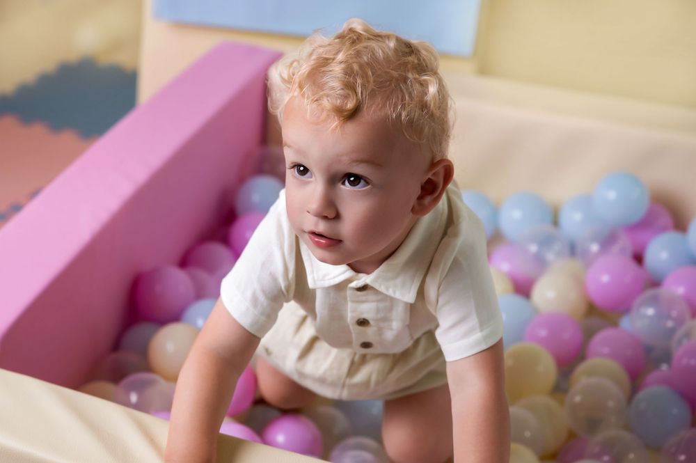 A toddler crawls near a pink ball pit filled with pastel-colored plastic balls in a playroom.