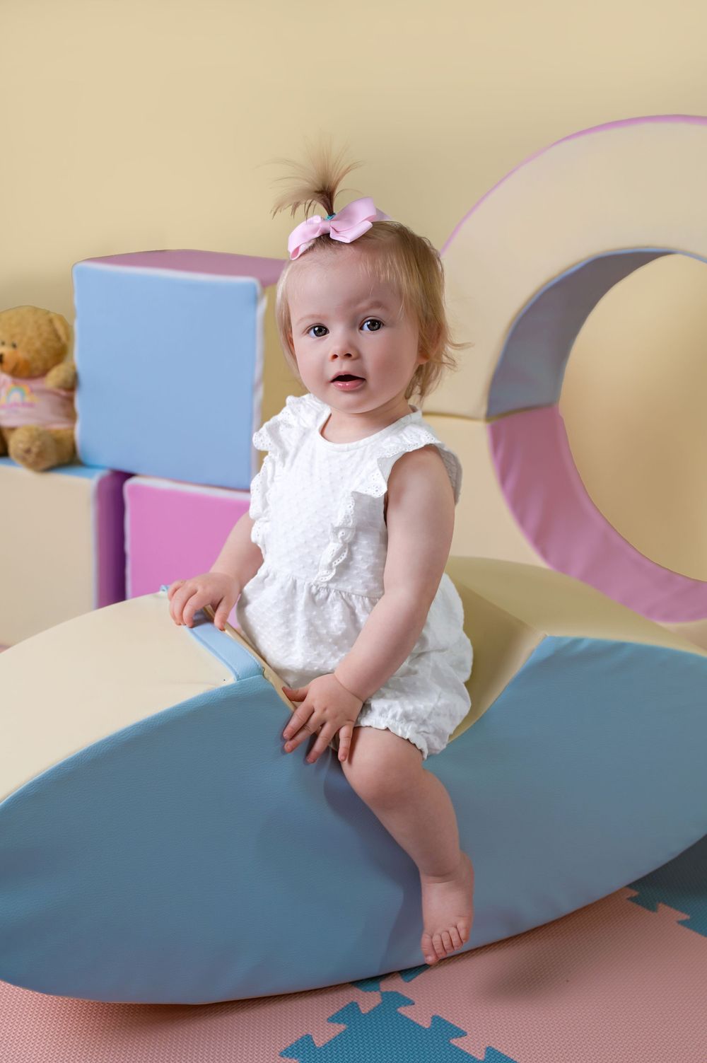 Baby in white outfit balanced on curved play furniture with colorful blocks behind.