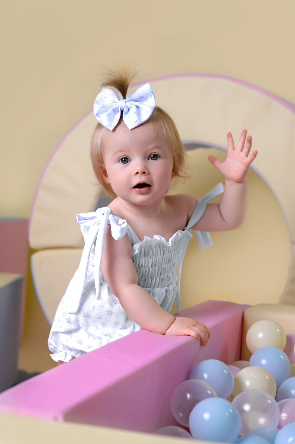 A small child wearing a white dress with a bow sits in a pink play area with pastel balloons.