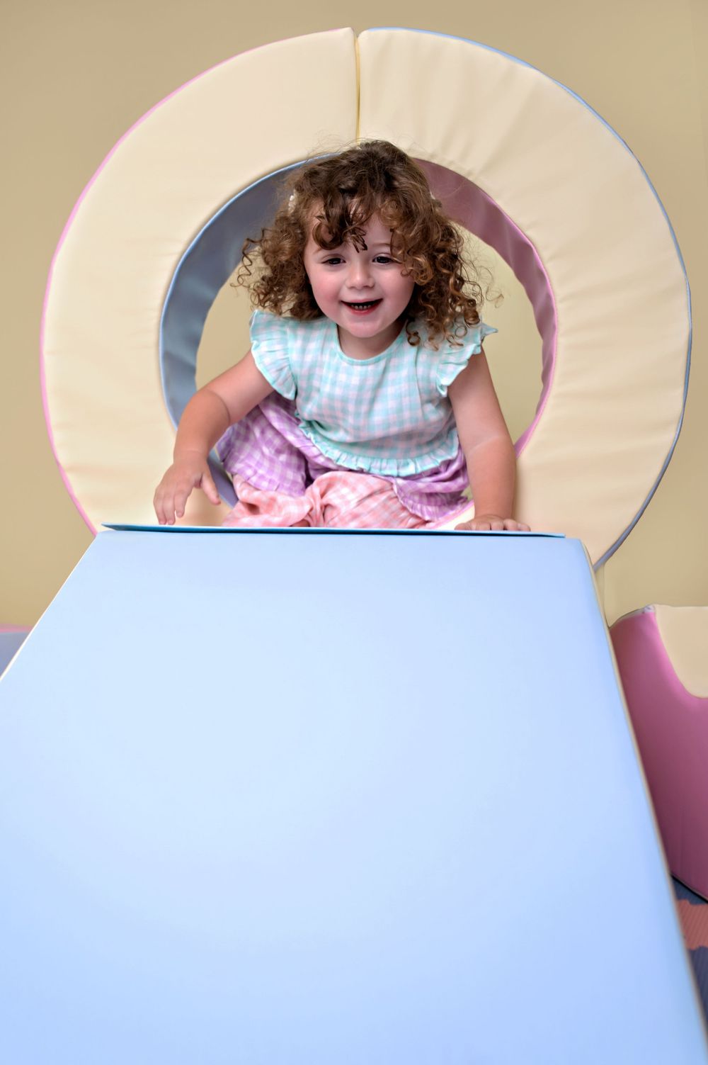 Young child with curly hair sitting and playing in a pastel-colored play area with an arch.